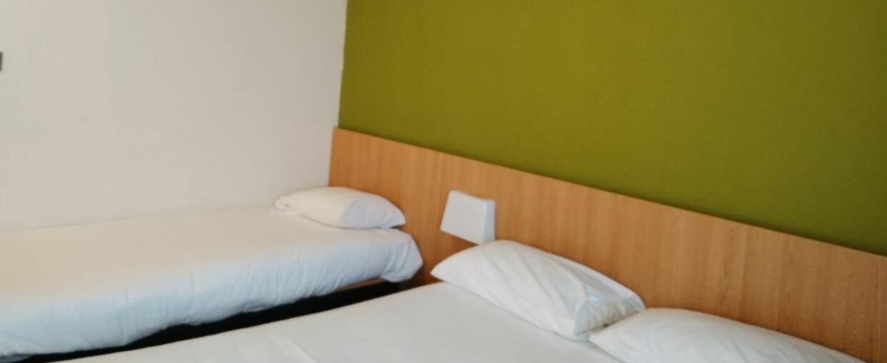 A room with two single beds, each with white bedding and pillows. The headboard is wooden, and the wall is painted green.