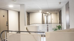 Un pasillo de hotel con barandilla de vidrio, paredes blancas y puertas que dan a las habitaciones.