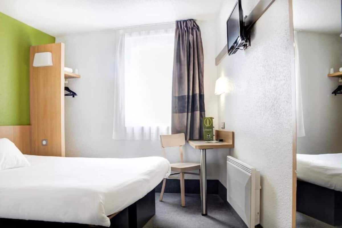 A hotel room with two beds, a small table with a chair, a TV mounted on the wall, and a window with curtains.