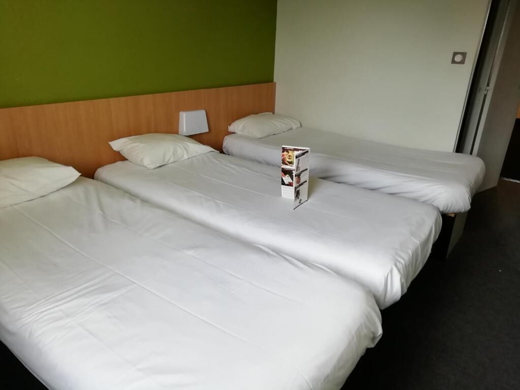 Quarto de hotel com três camas individuais, cada uma com roupa de cama branca e cabeceira de madeira. Uma pequena mesa com uma revista está colocada entre as camas.