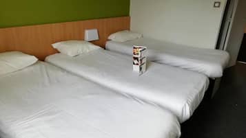 Quarto de hotel com três camas individuais, cada uma com roupa de cama branca e cabeceira de madeira. Uma pequena mesa com uma revista está colocada entre as camas.