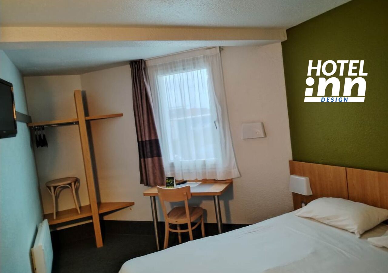 A hotel room with a bed, a desk with a chair, a window with curtains, and a wall-mounted shelf.