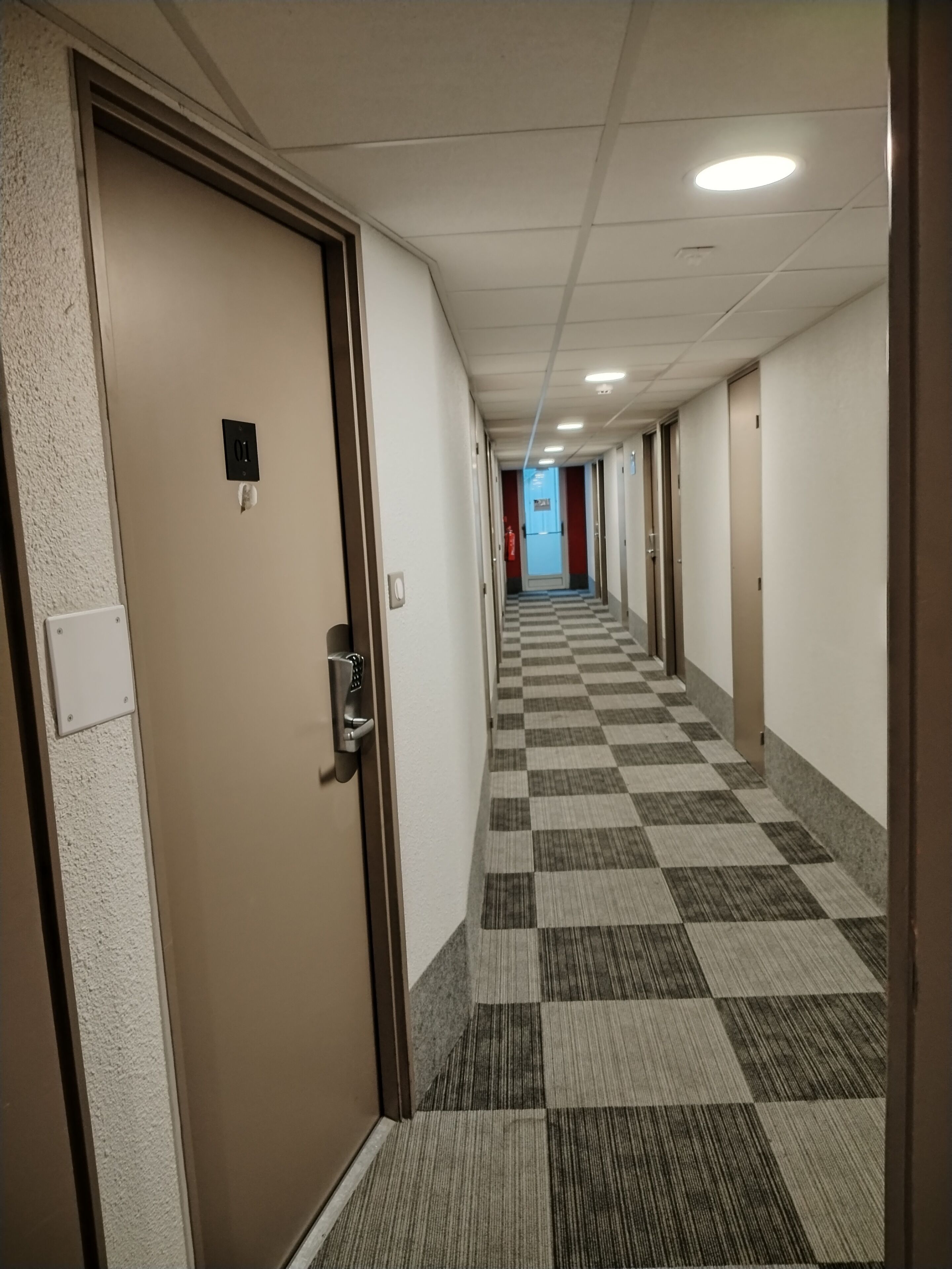 Ein Hotelgang mit Schachbrettmuster-Teppich, Türen links und einem Feuerlöscher an der Wand.