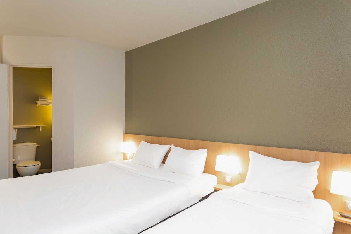 A hotel room with two beds, a wooden headboard, bedside lamps, and a small bathroom visible through an open door.