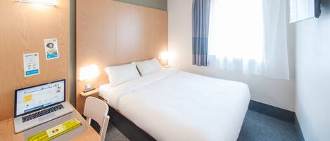 Una habitación de hotel con una cama, un escritorio con una computadora portátil, una lámpara y un televisor instalado en la pared.