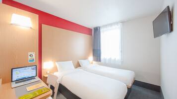 A hotel room with two beds, a desk with a laptop, a flat-screen TV mounted on the wall, and a window with curtains.