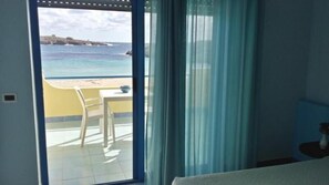A room with a view of a beach and ocean, a chair, a table, and a window with curtains. - Hotel Baia Turchese (Lampedusa)