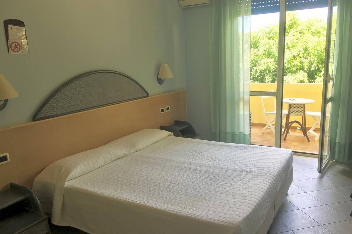 A hotel room with a bed, a nightstand, a lamp, and a view of a balcony with outdoor furniture.