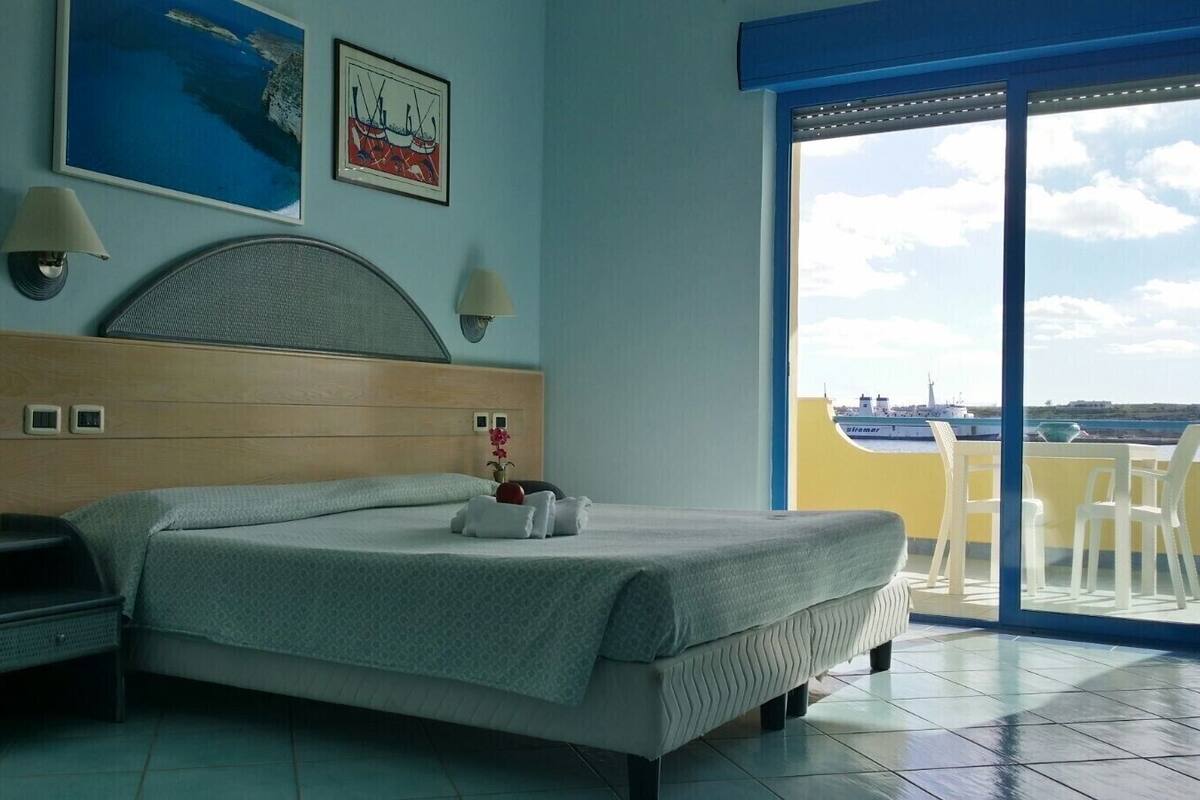 A hotel room with a bed, a desk with a telephone, two wall-mounted lamps, and a large window with a view of the sea and boats.
