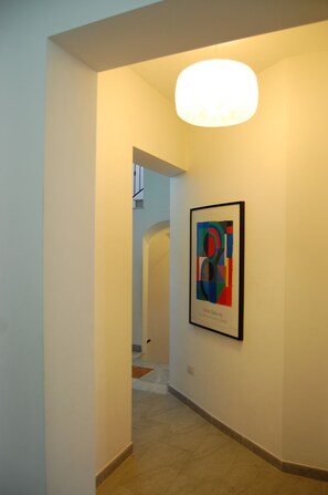 A hallway with a framed abstract painting on the wall, a staircase in the background, and a light fixture above. - Helios B&B (Castellammare del Golfo)