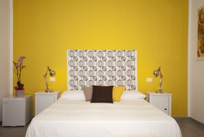 A hotel room with a yellow wall, a patterned headboard, white bedding, and two bedside tables with lamps. - Helios B&B (Castellammare del Golfo)