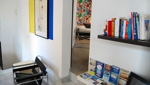 A room with a desk, a chair, a bed, a colorful wall, and a shelf with books and maps.
