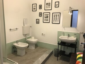 A bathroom with a toilet, bidet, sink, and a small shelf. There are framed pictures on the wall and a mirror above the sink. - Helios B&B (Castellammare del Golfo)