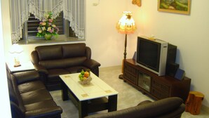 Un salon avec un canapé, une table basse sur laquelle on a disposé des fruits, un téléviseur posé sur un meuble en bois et une fenêtre ornée de rideaux en dentelle.
