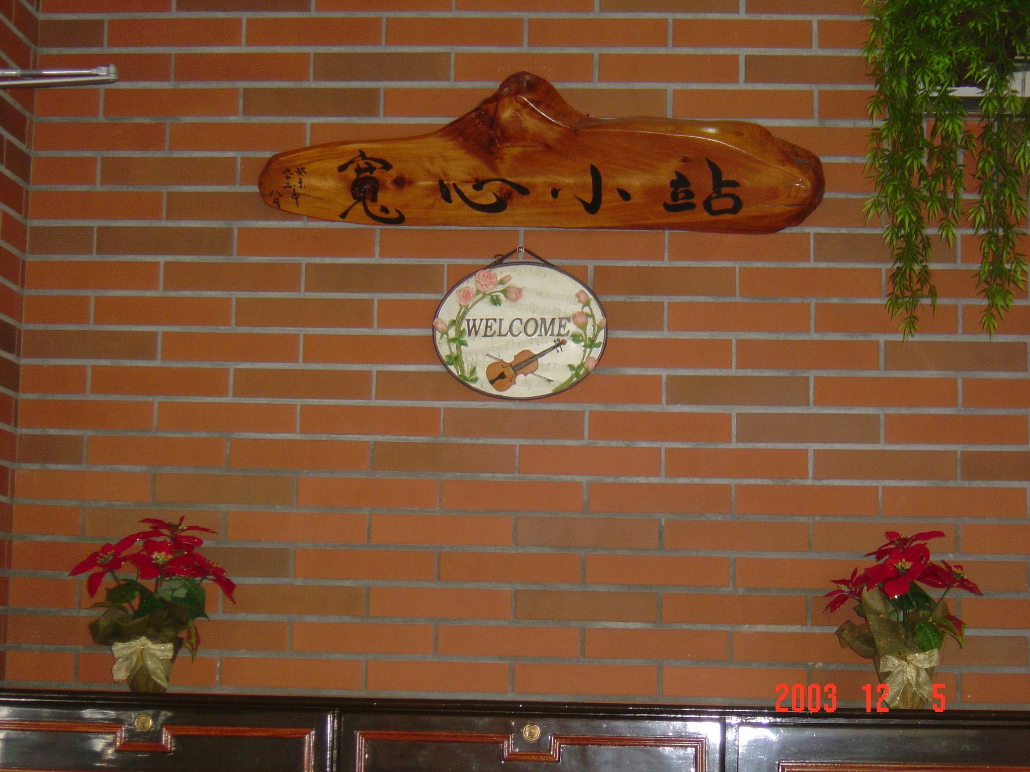 A wooden welcome sign with Japanese characters, a 'WELCOME' sign with a guitar, and two potted plants on a brick wall.