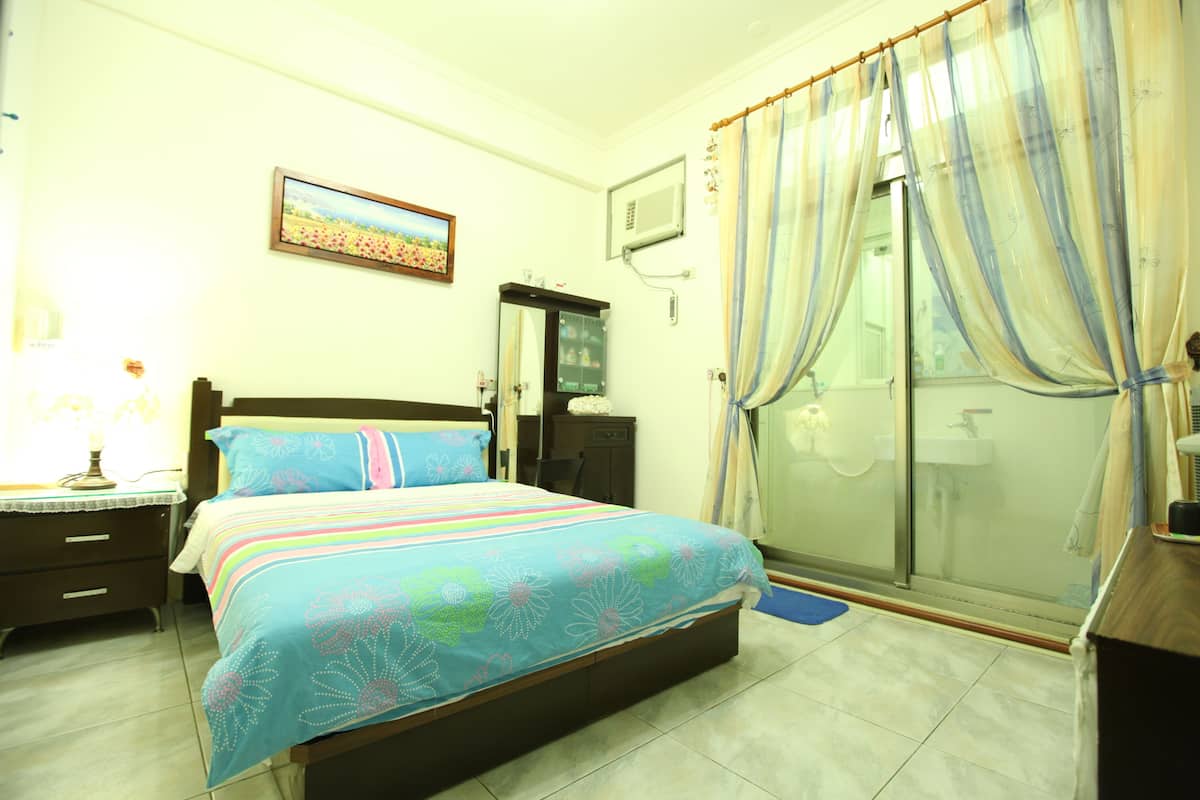 Un dormitorio con cama, mesita de noche, cómoda, espejo, ventana con cortinas y zona de ducha.