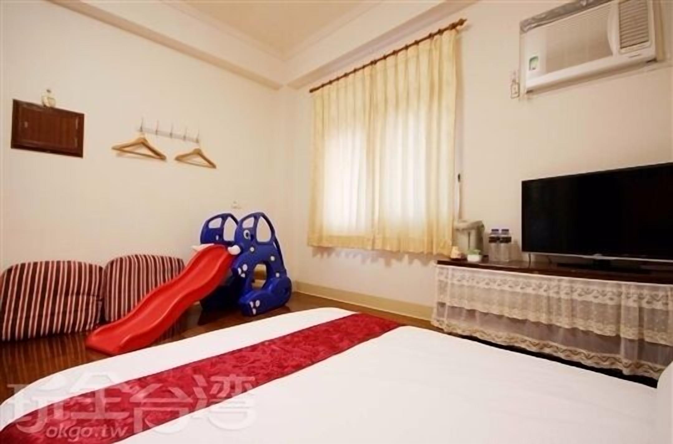 Quarto de hotel com cama, televisão, janela com cortinas, ar-condicionado fixado na parede e uma pequena mesa com objetos.
