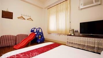 A hotel room with a bed, a television, a window with curtains, a wall-mounted air conditioner, and a small table with items.