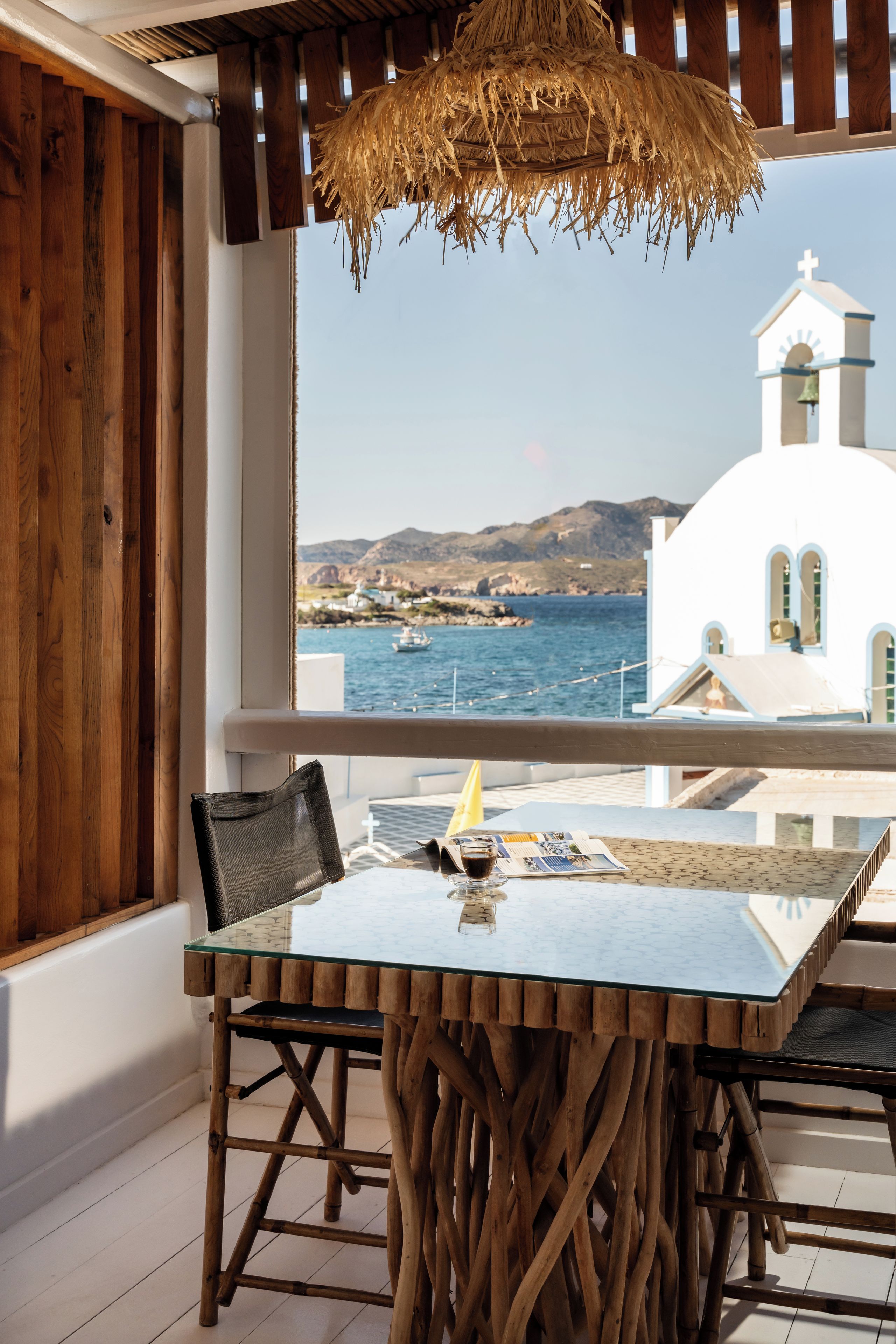 Una mesa con cubierta de vidrio y patas de madera, una silla, una lámpara colgante y vistas de un edificio blanco con un campanario junto al mar.