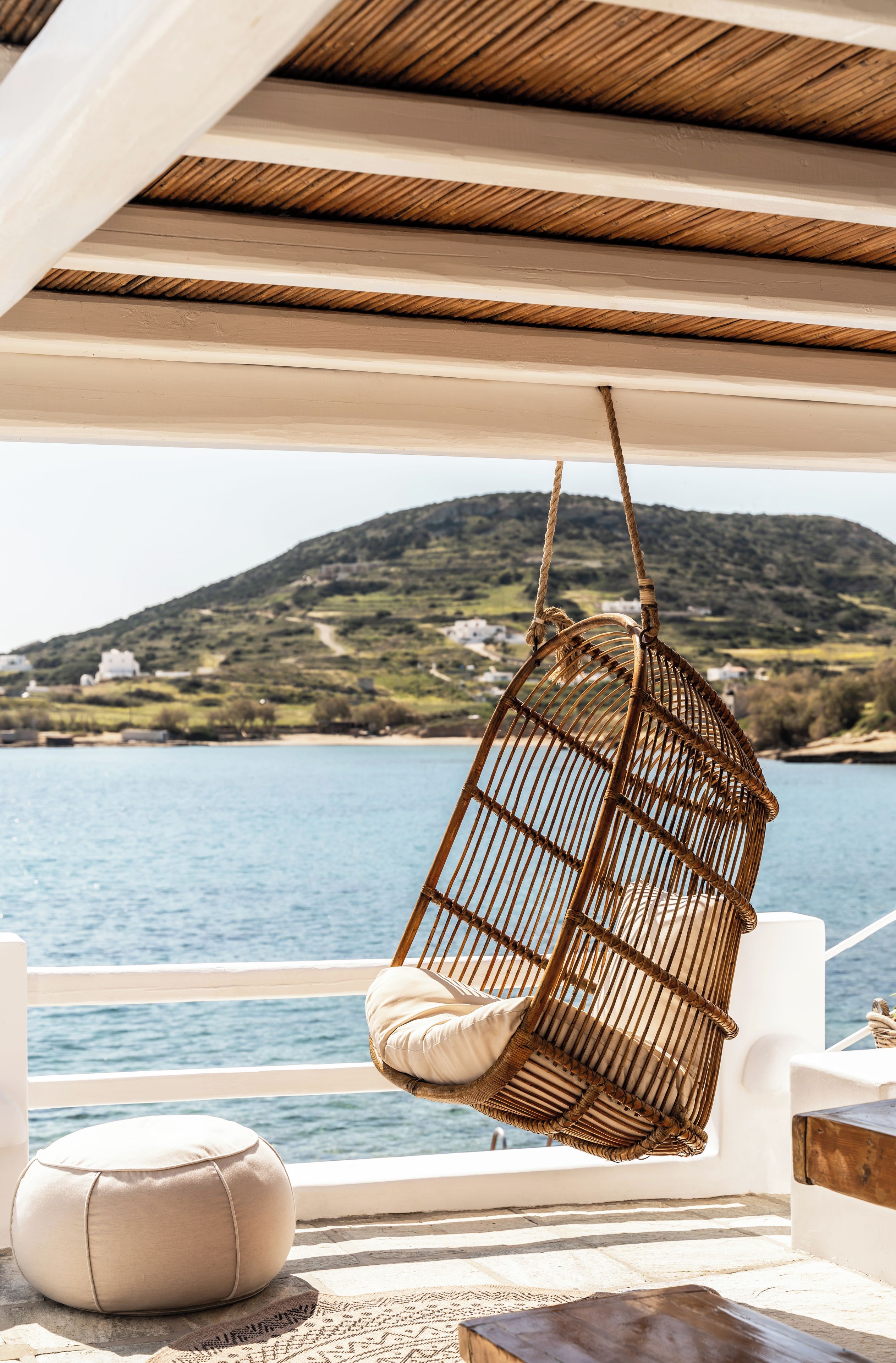 Una silla colgante de mimbre con cojines, un reposapiés blanco y una mesa de madera en un balcón con vistas a un cuerpo de agua y un paisaje montañoso.