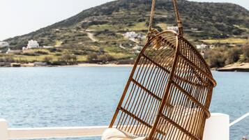 Una silla colgante de mimbre con cojines, un reposapiés blanco y una mesa de madera en un balcón con vistas a un cuerpo de agua y un paisaje montañoso.