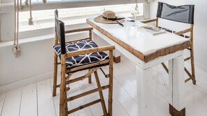 A room with a wooden table and chairs, a woven ceiling light, a view of the sea, and a white wall with a woven wall hanging. - Captain Zeppos (Milos)
