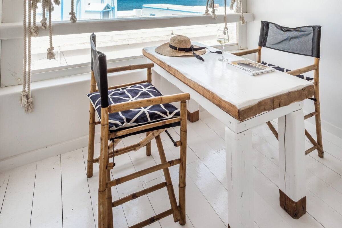 Ein Zimmer mit Holztisch und -stühlen, einer gewebten Deckenleuchte, Blick auf das Meer und einer weißen Wand mit Wandbehang.