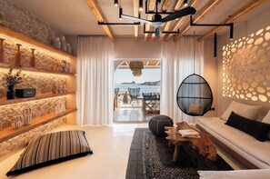 A modern living room with a stone wall, a wooden ceiling, a white sofa, and a view of the sea. - Captain Zeppos (Milos)