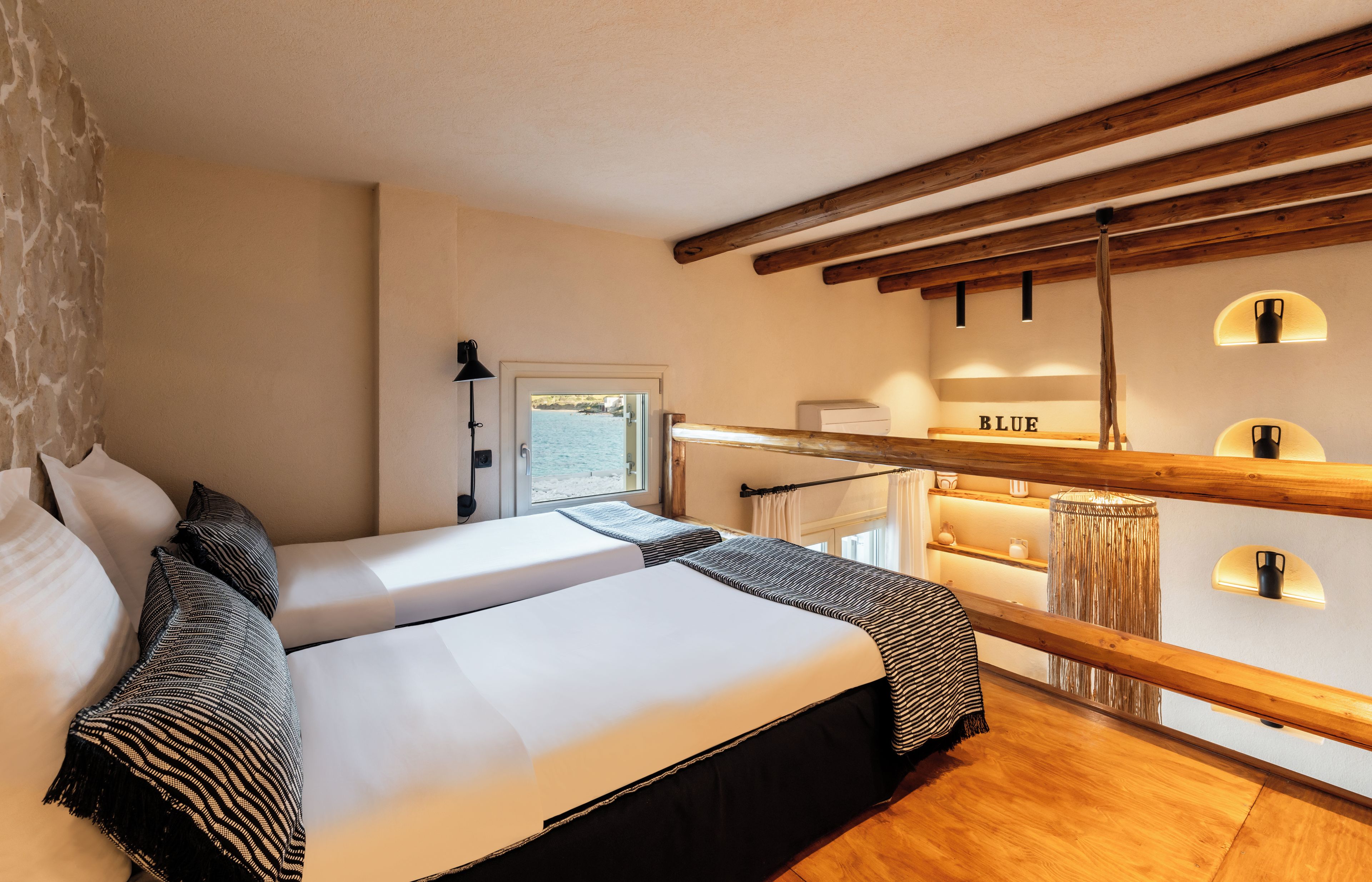 Cama doble con sábanas blancas y almohadas a rayas blancas y negras, techo de madera y vista al mar a través de una puerta.