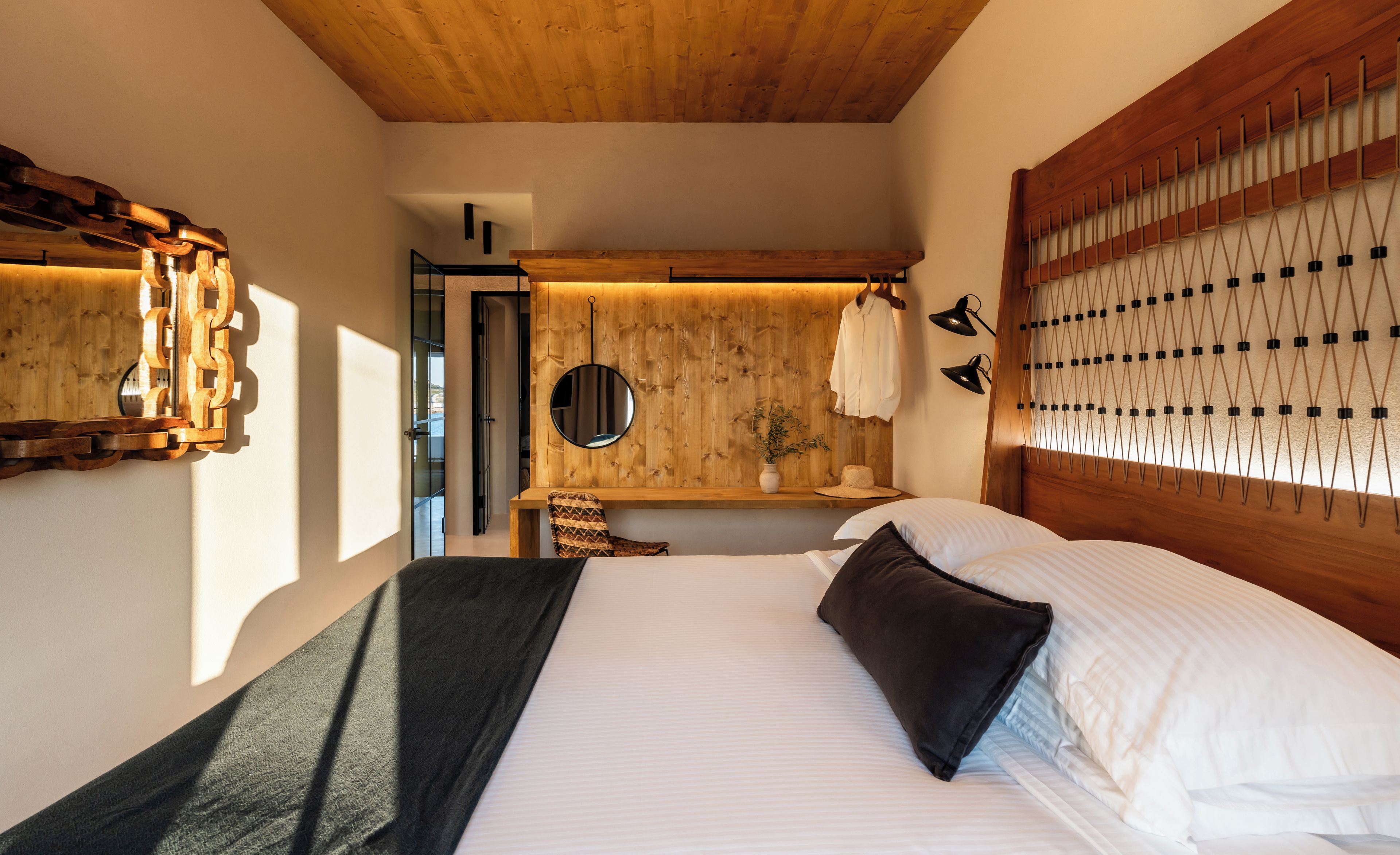 Un dormitorio con techo de madera, una cama con ropa de cama blanca, una mesita de noche con espejo y una lámpara de pared.