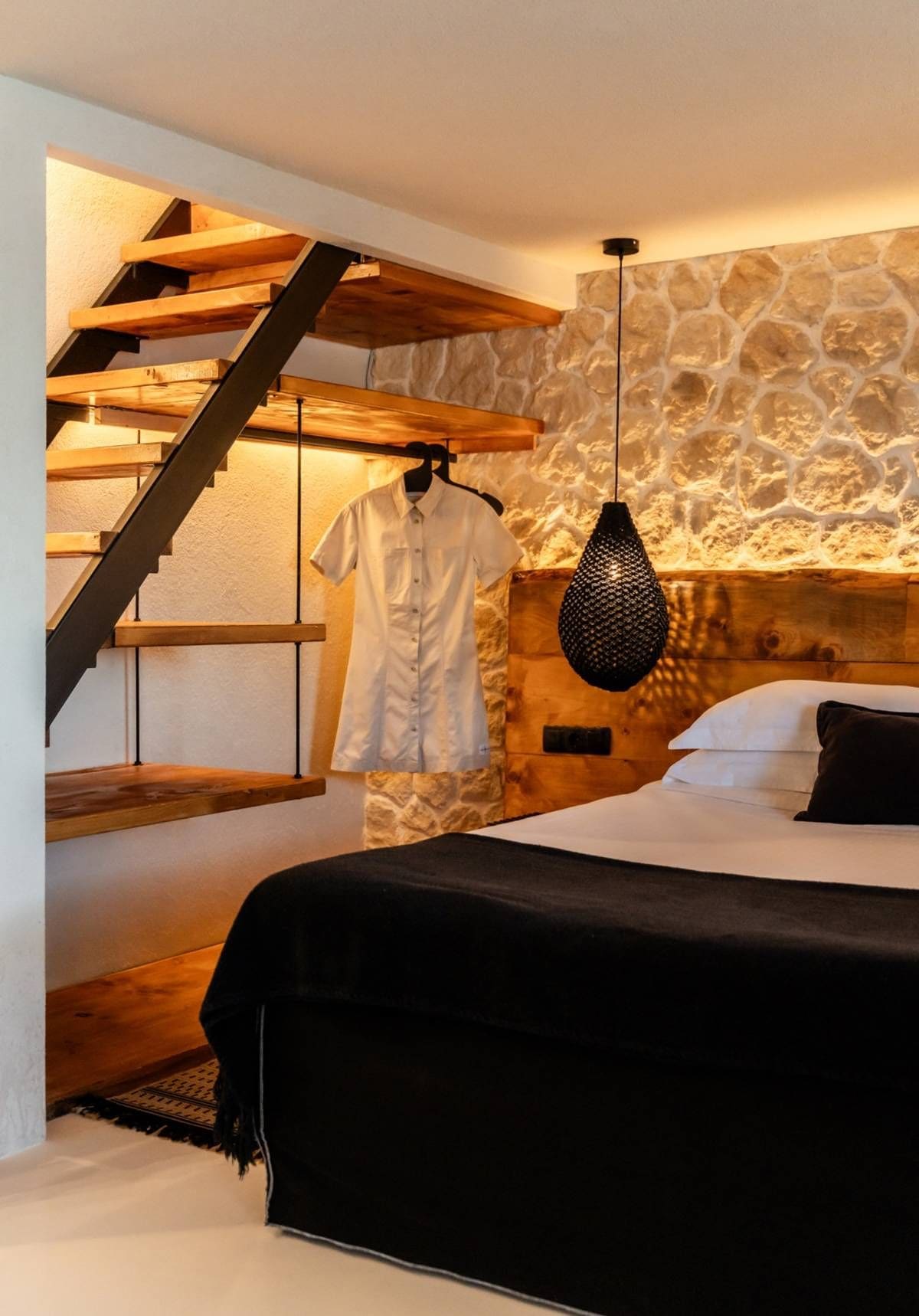 Une chambre avec un lit, un luminaire suspendu, un escalier en bois et un mur en pierre.