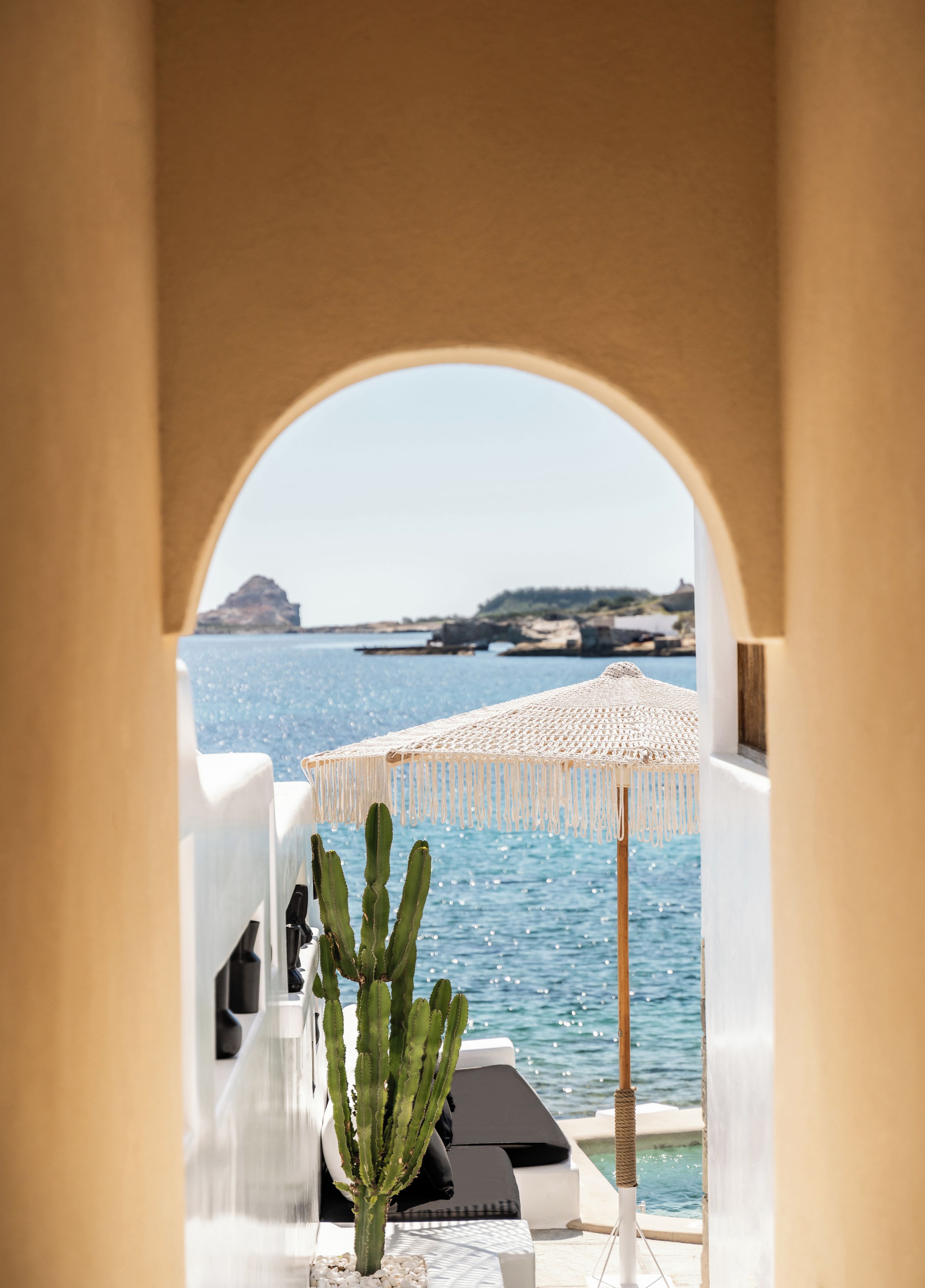 A view through an archway to a pool area with a cactus plant, lounge chairs, and a beach in the background.