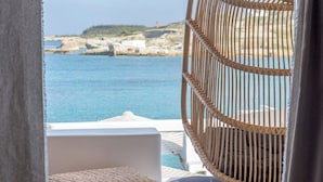 A wicker hanging chair overlooking a coastal view with a white cushioned bench and woven rugs. - Captain Zeppos (Milos)
