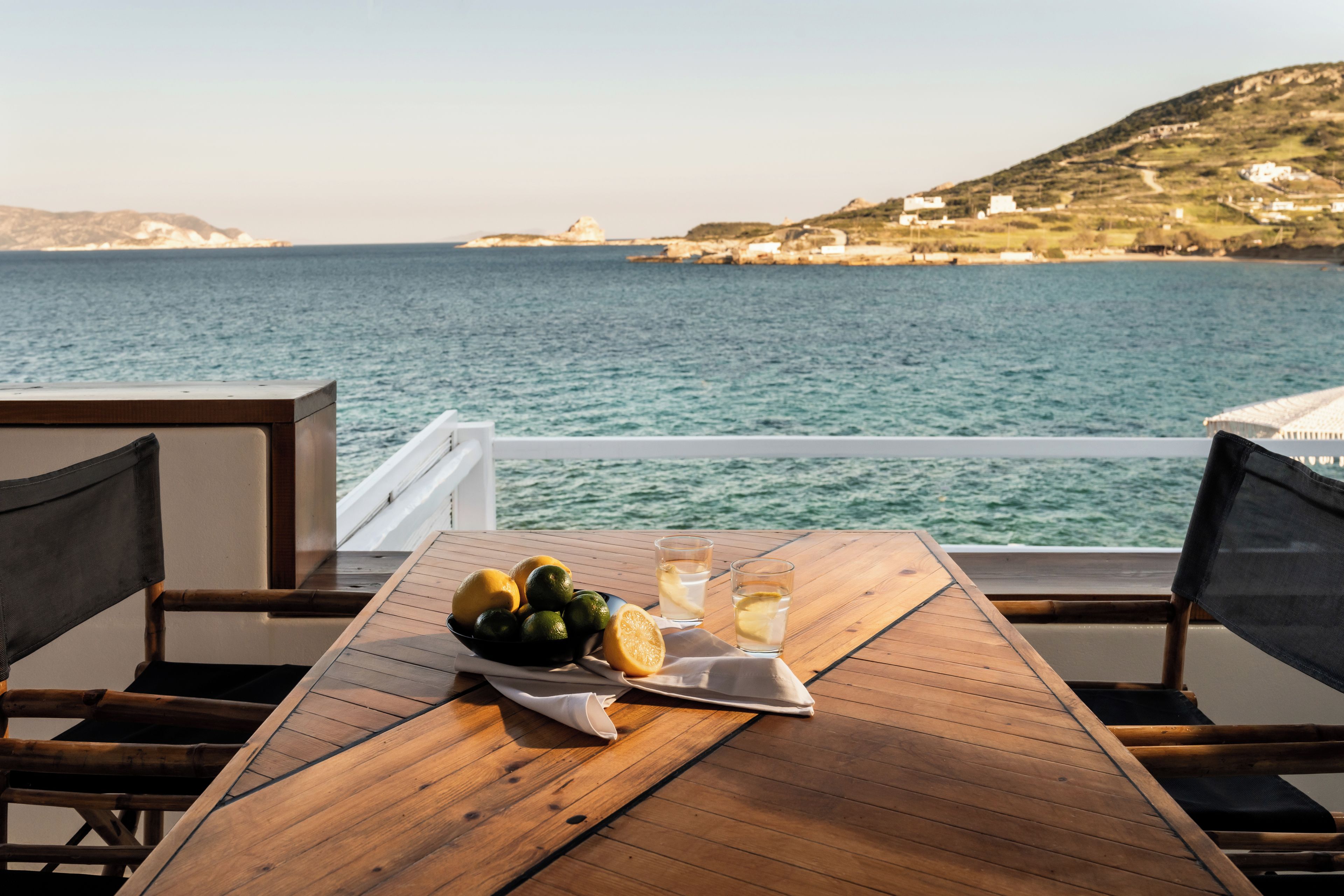 Una mesa de madera con un bol de fruta y vasos de agua, con vista a una masa de agua y una costa lejana.