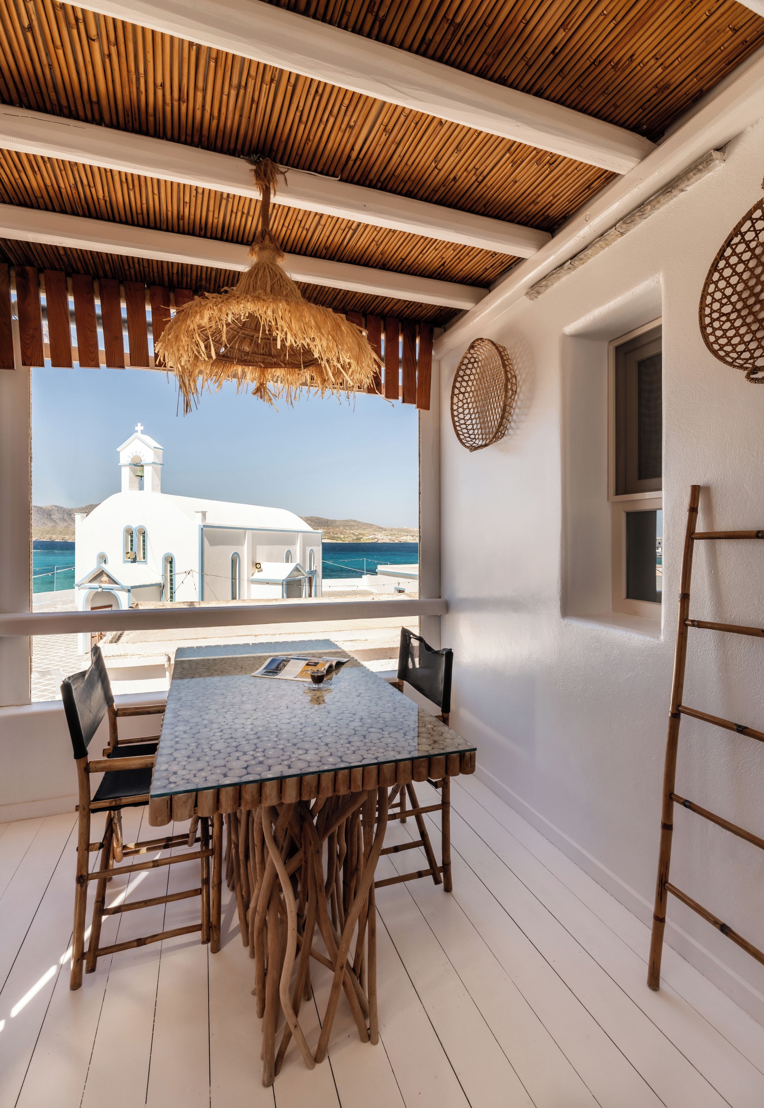 Un espacio de comedor al aire libre cubierto con una mesa, sillas, una lámpara colgante y vistas al mar y un edificio blanco.