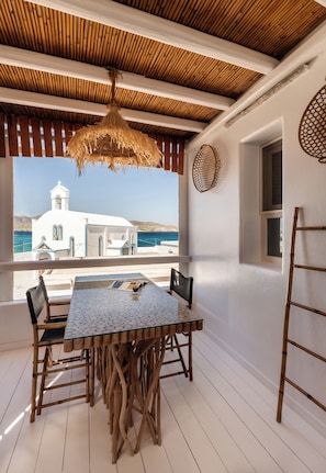 A covered outdoor dining area with a table and chairs, a hanging light fixture, and a view of the sea and a white building. - Captain Zeppos (Milos)