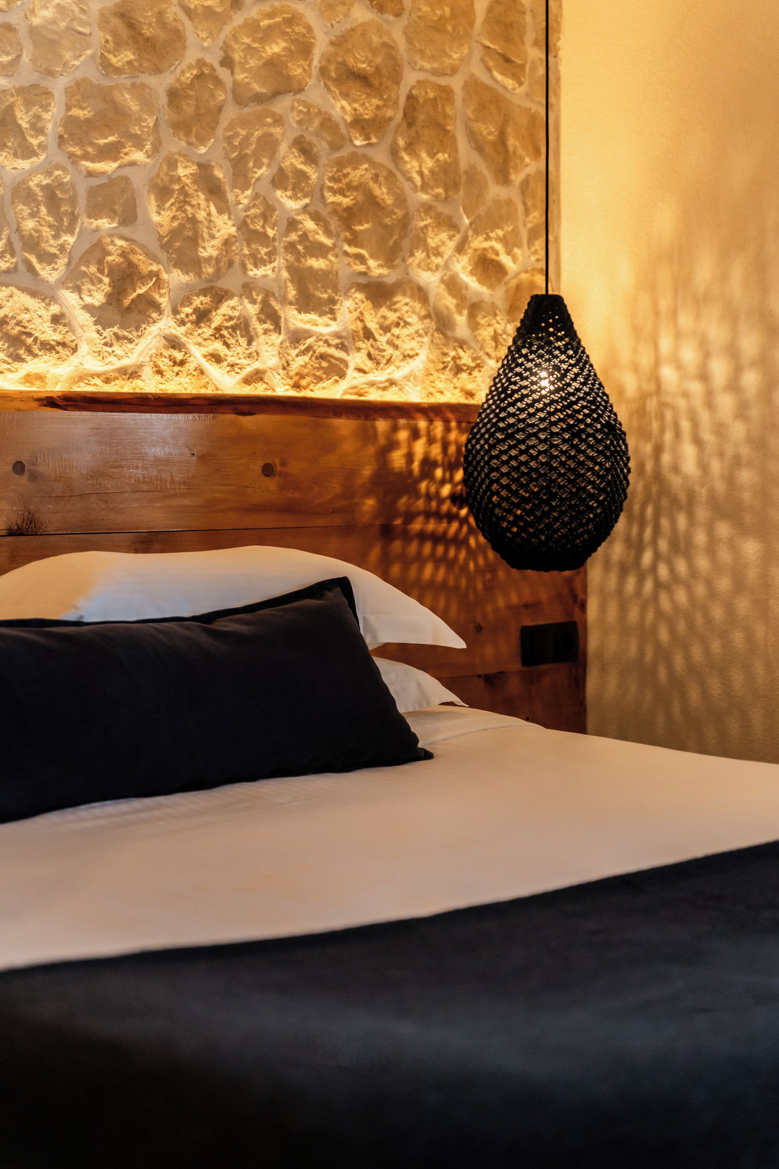 Una cama con ropa de cama blanca y negra, cabecero de madera y pared de piedra texturizada al fondo.