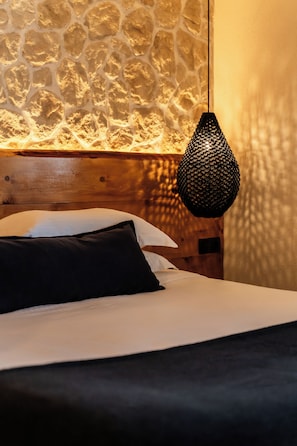 A bed with white and black bedding, a wooden headboard, and a textured stone wall behind. - Captain Zeppos (Milos)
