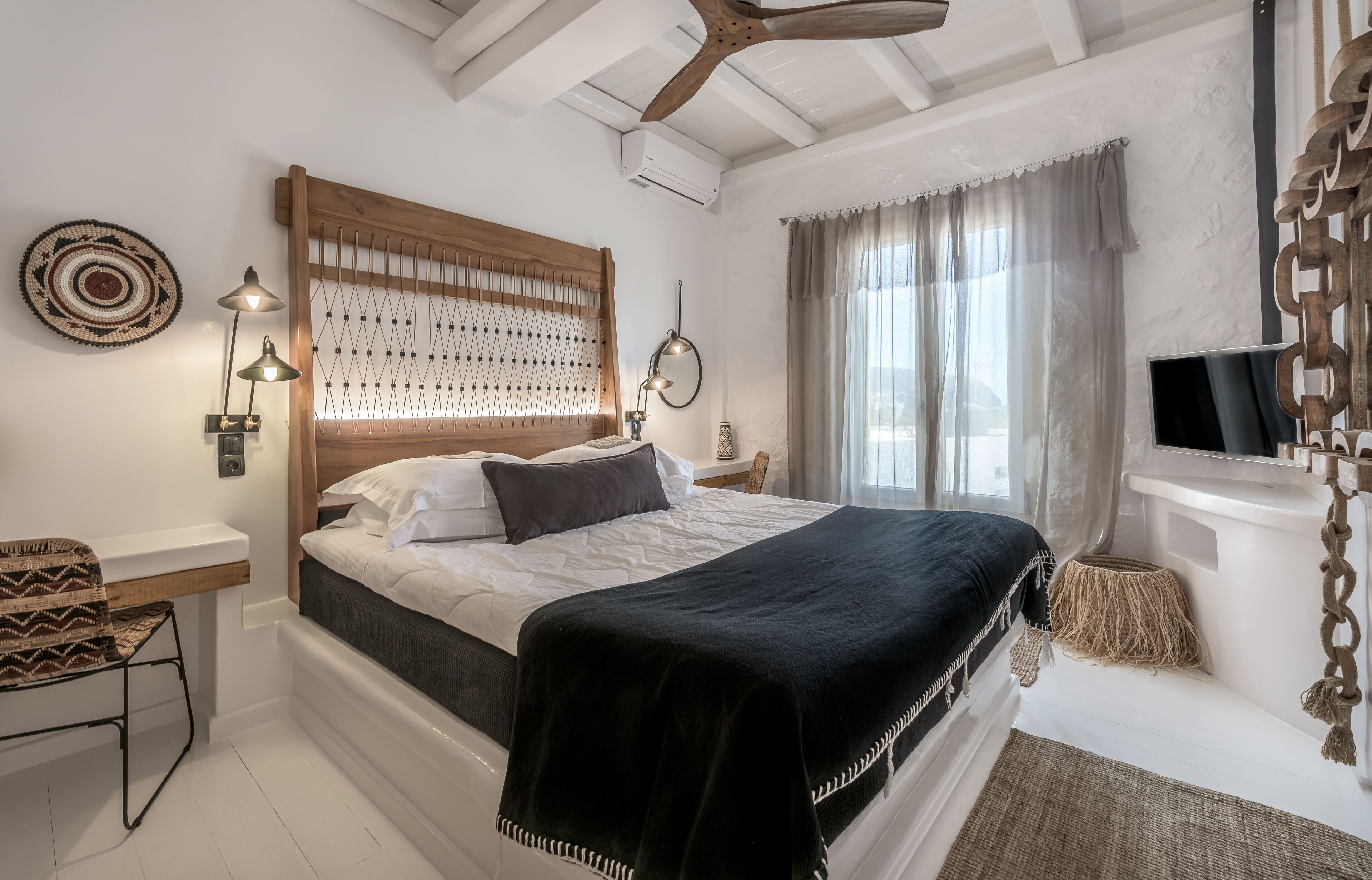 Un dormitorio con cabecera de madera, cama con ropa blanca y un plaid negro, lámpara de noche, televisor y ventana con cortinas.