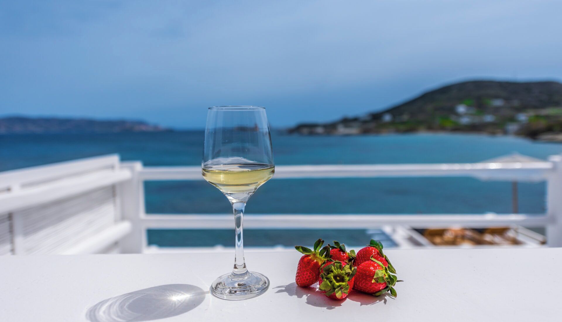 A glass of white wine on a white table with a view of the sea and a white railing.