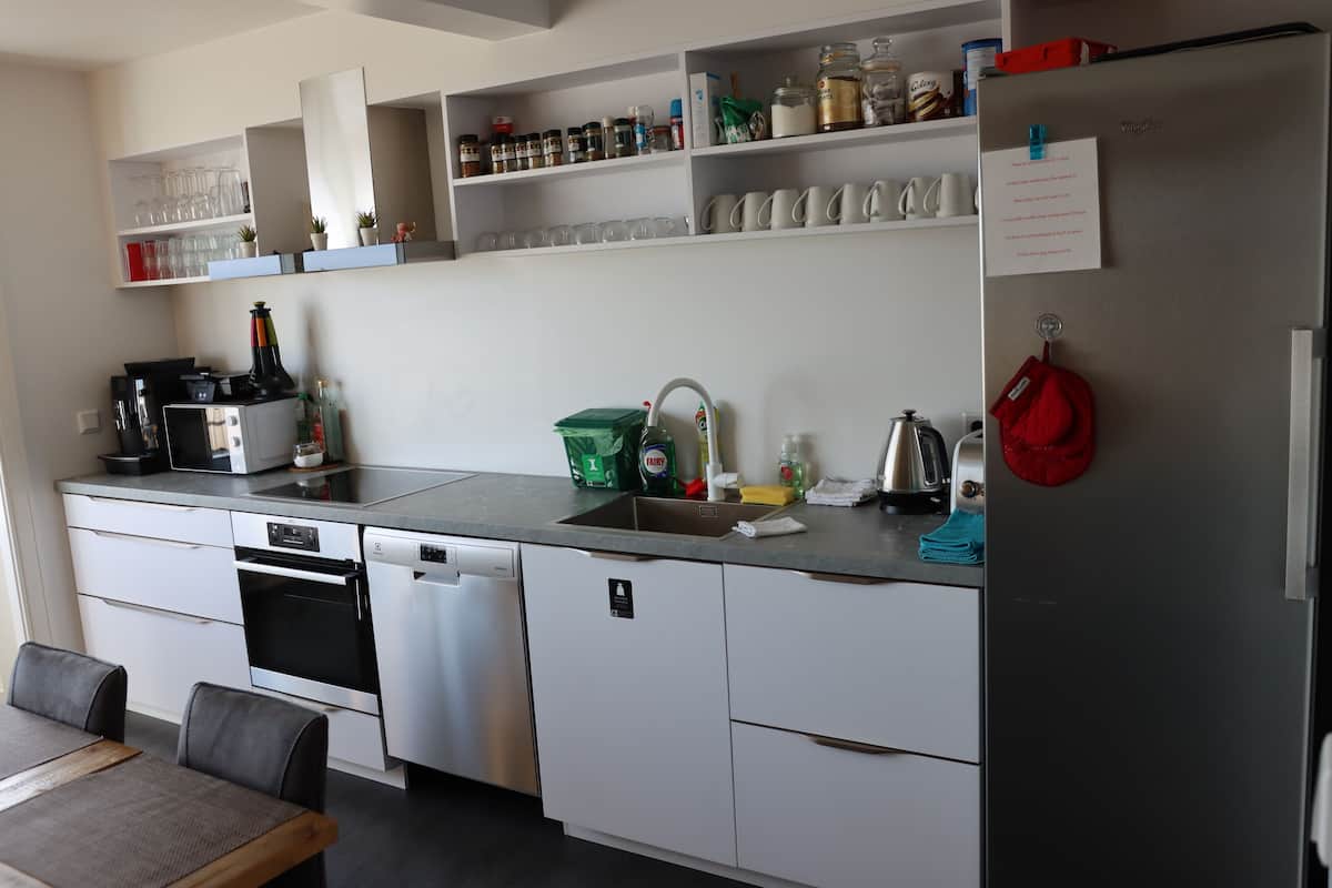 A modern kitchen with white cabinets, a stainless steel dishwasher, a built-in oven, and a refrigerator. There are shelves above the counter holding various kitchen items.