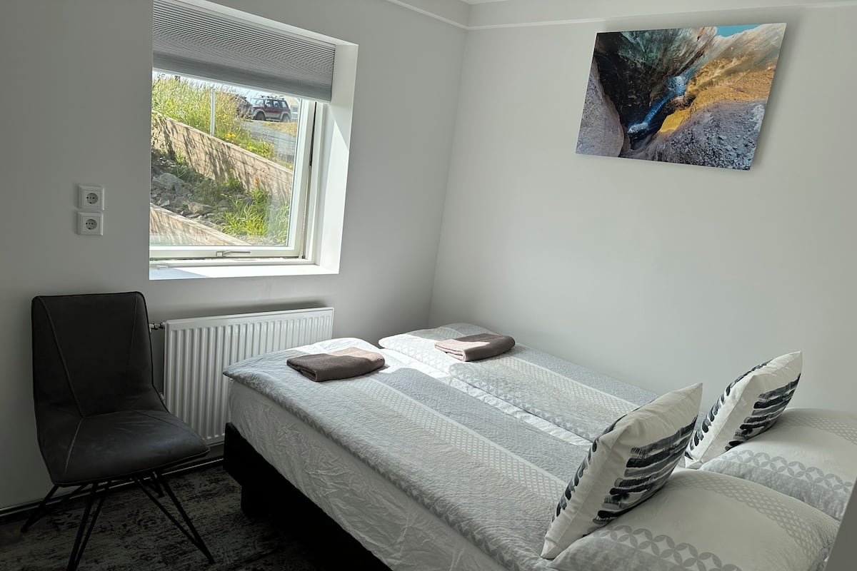 A small, clean room with a bed, a chair, a radiator, a window with a view of a vehicle, and a painting on the wall.