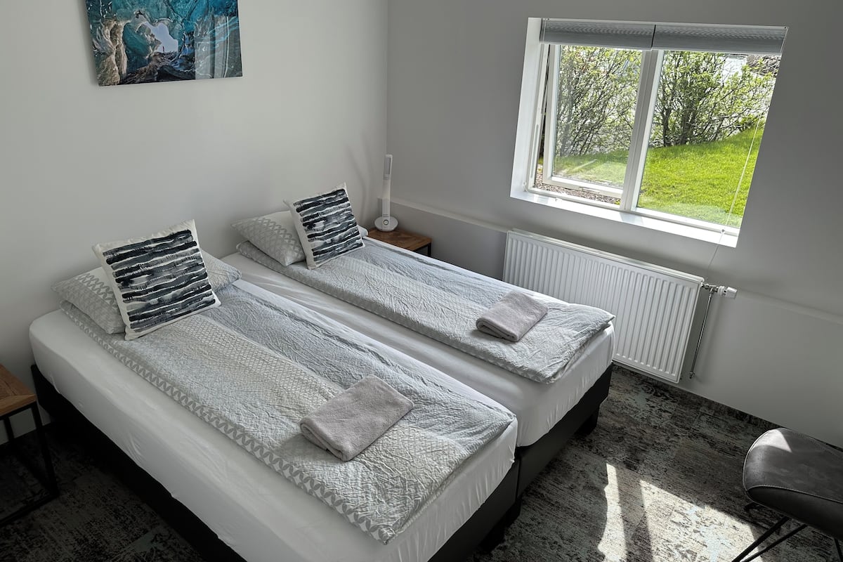 A bedroom with two beds, a window with a view of greenery, a bedside table with a lamp, and a painting on the wall.