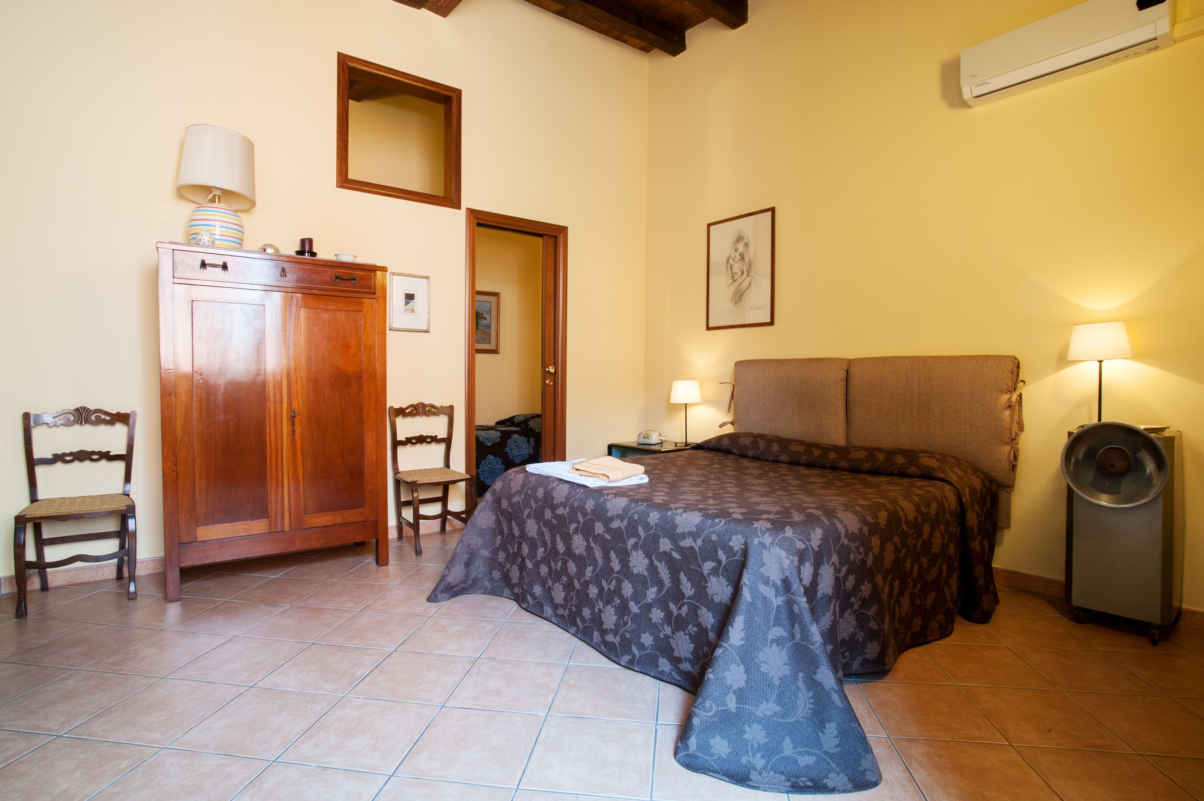 A bedroom with a bed, a wooden wardrobe, a chair, a bedside table with a lamp, and a wall-mounted air conditioner.