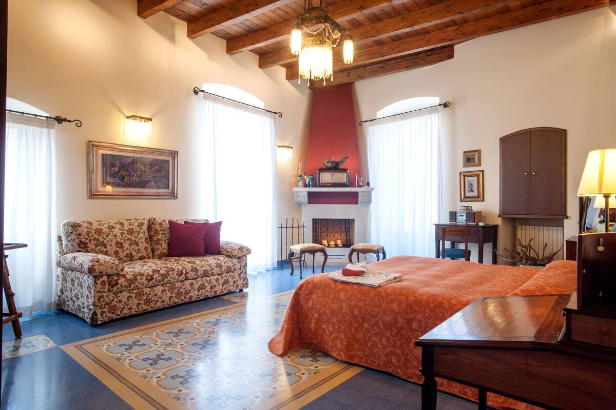 A hotel room with a bed, a sofa, a fireplace, and a chandelier.