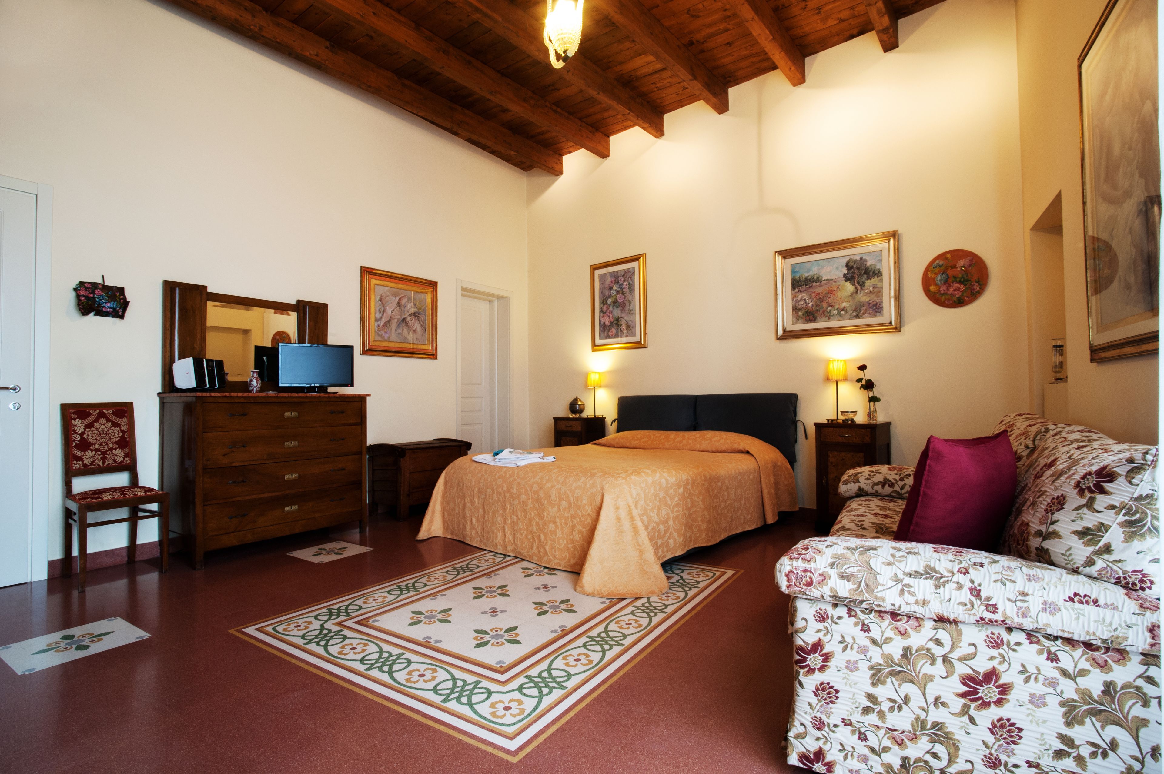 A bedroom with a bed, a dresser, a chair, a sofa, a TV, and framed artwork on the walls.
