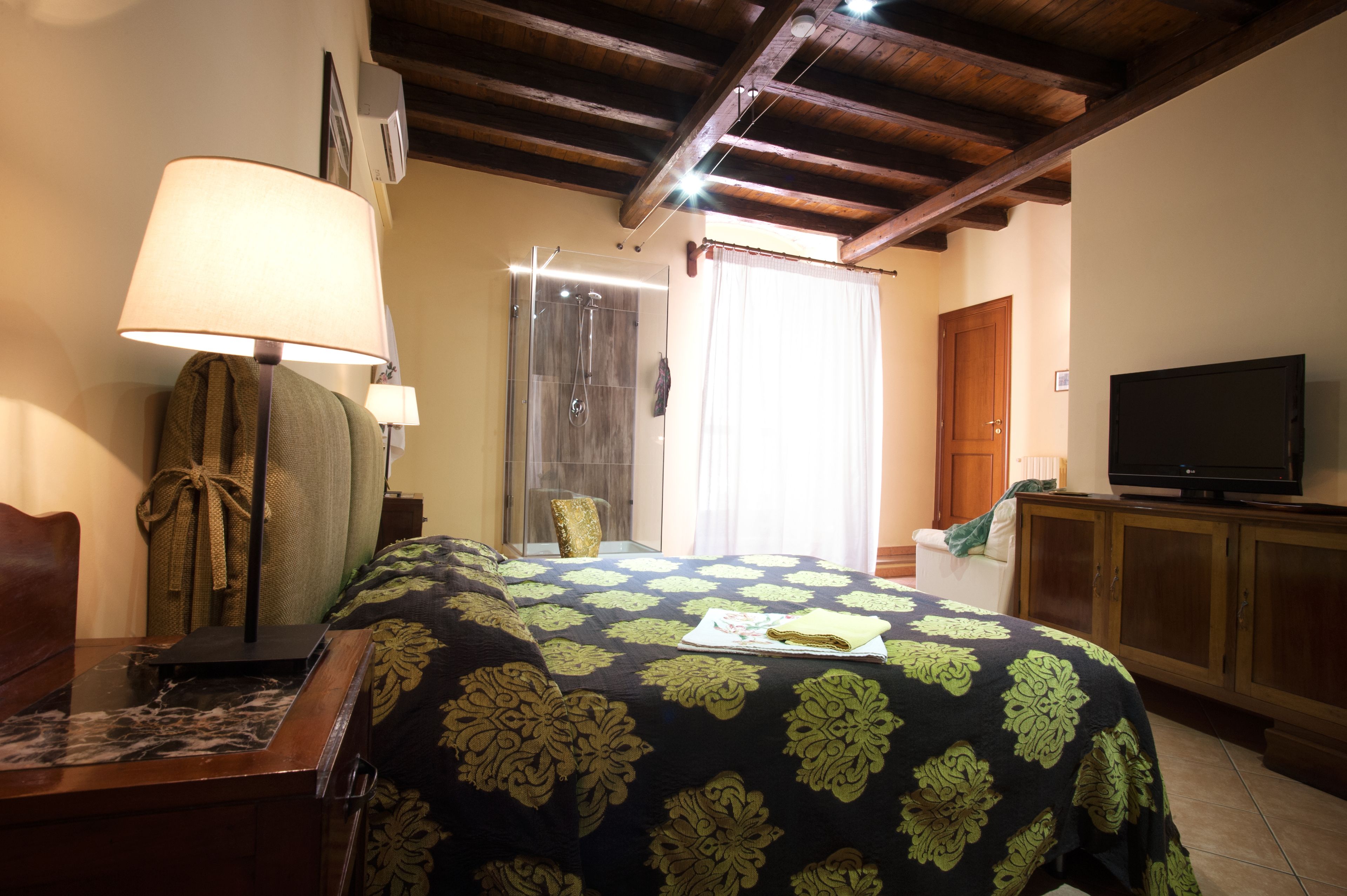 A hotel room with a bed, a TV, a desk with a lamp, a shower, and a wooden ceiling.