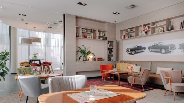 Een moderne woonkamer met een eethoek, een boekenkast en een groot kunstwerk op de muur, waarop oldtimers afgebeeld zijn.