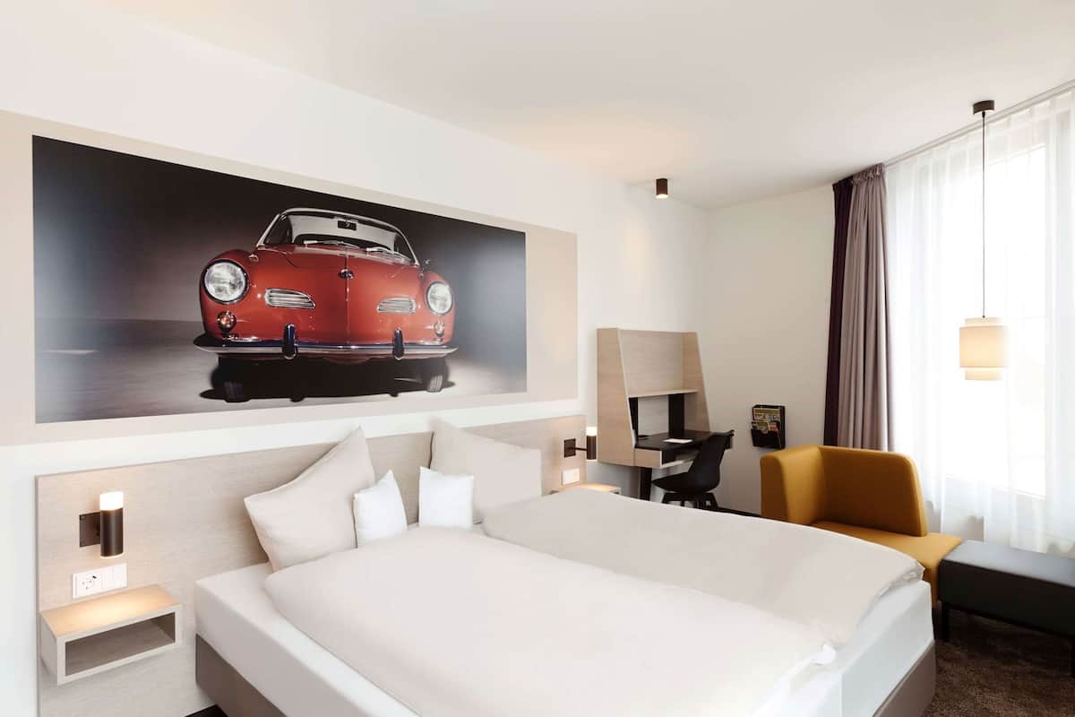 Habitación de hotel con una cama grande, un escritorio, una silla, una lámpara de escritorio, un televisor montado en la pared y una ventana con cortinas.