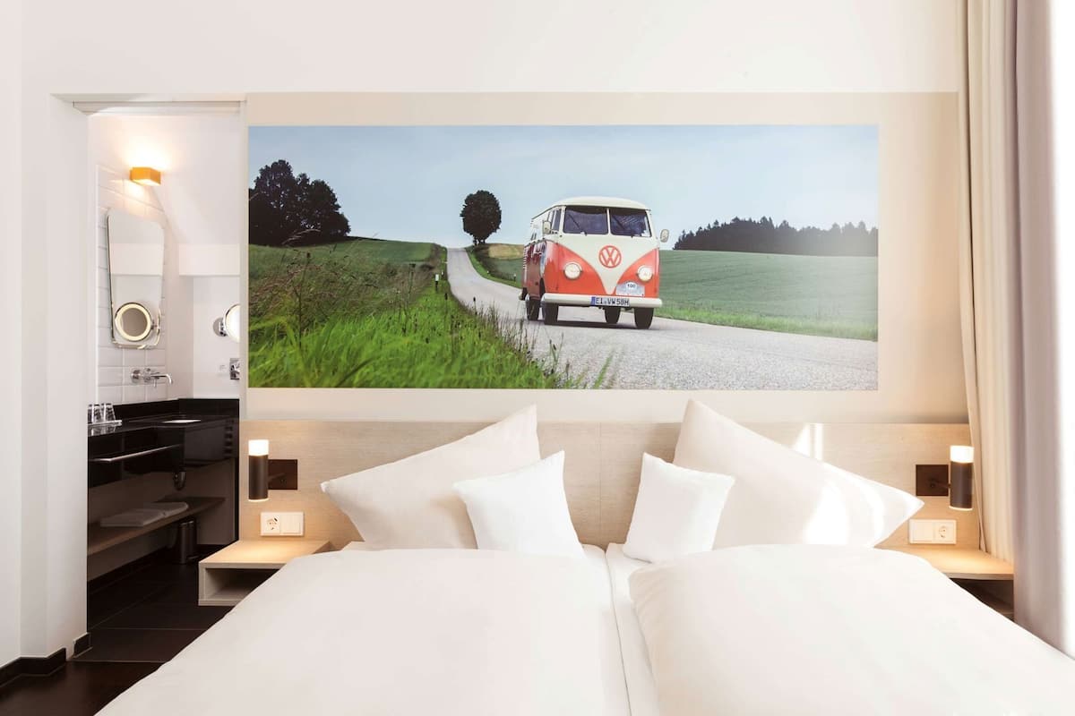 Un dormitorio con una cama grande, una mesita de noche con lámpara, un espejo colgado en la pared y un cuadro de una furgoneta en una carretera.